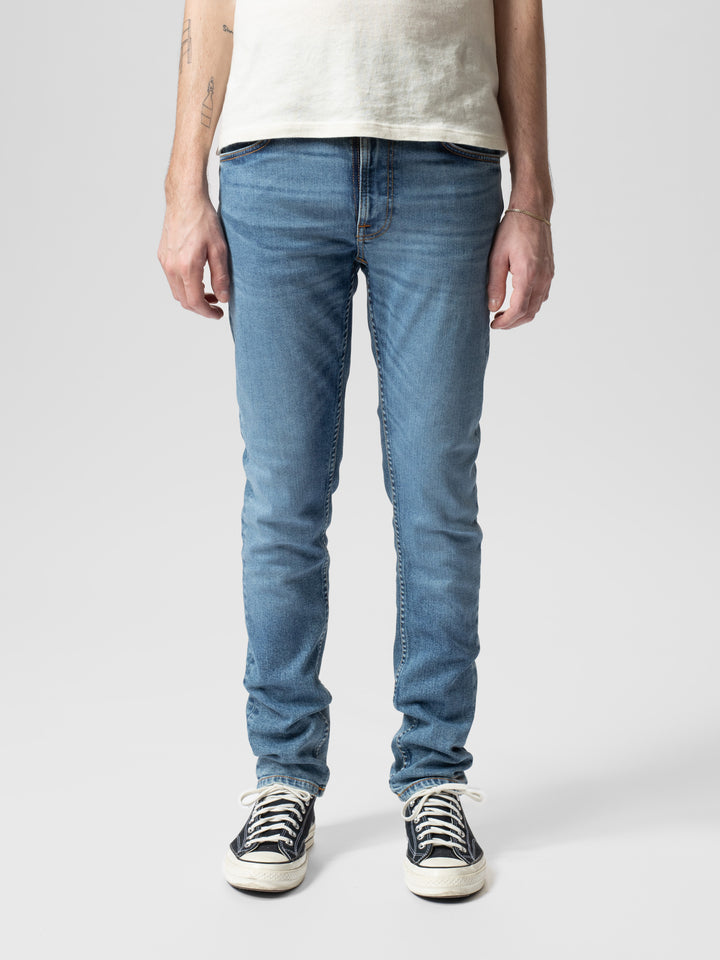 Person wearing light blue Nudie jeans and white sneakers on a plain background